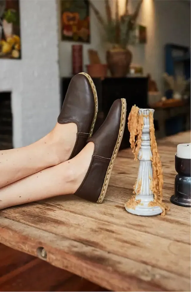 Feet in dark brown leather slip-on shoes propped on a rustic wooden table beside a candlestick